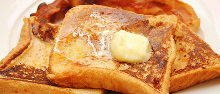 Two slices of French toast from The Brick Menu, topped with melting butter, crispy bacon behind.